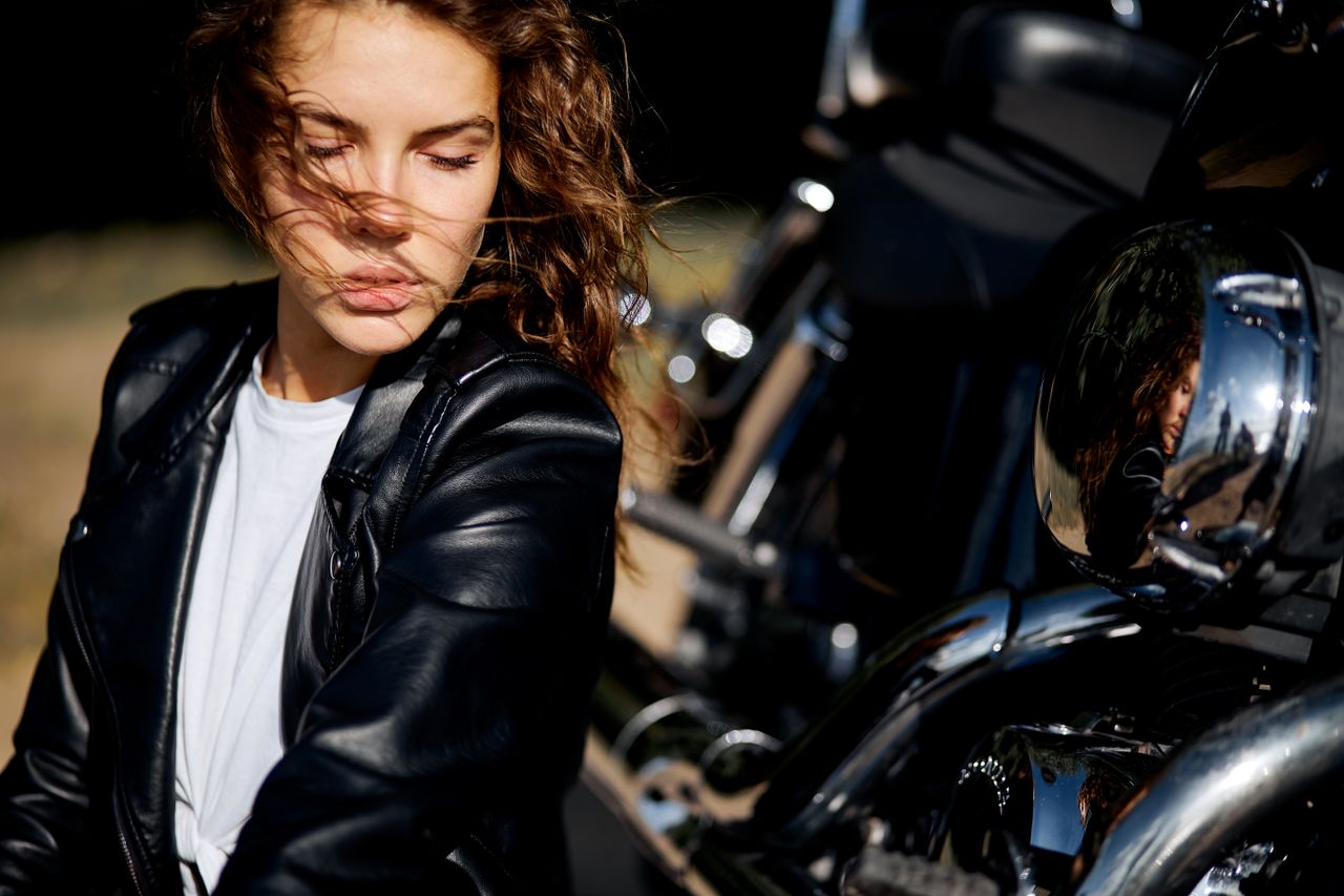 Woman in leather jacket posing beside a shiny motorcycle outdoors.