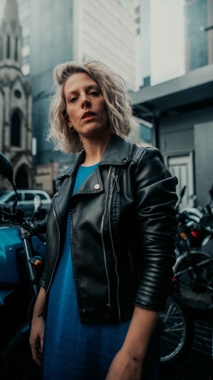 Blonde woman wearing a leather jacket standing on a city street surrounded by motorcycles.