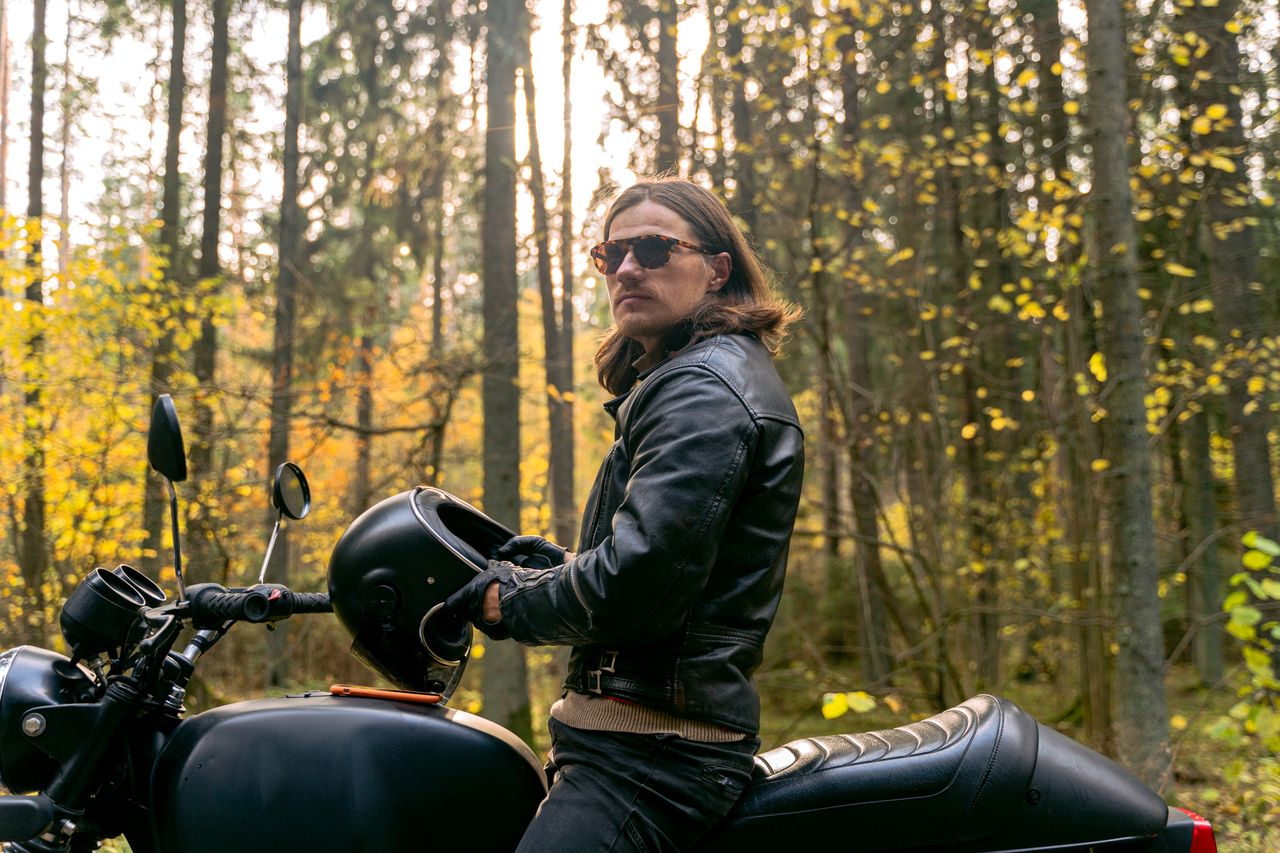 Man in black leather jacket with a motorcycle helmet in an autumn forest setting.